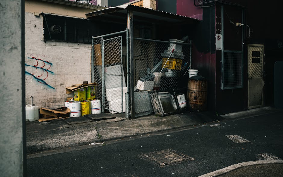 A small outdoor storage area situated on a paved urban street, enclosed by a metal chain-link fence, contains various discarded items related to rubbish clearance. Visible objects include multiple large white plastic buckets and yellow containers stacked near the base of the fence, alongside flattened cardboard boxes. Adjacent to these, there are several metal trays, a wooden barrel, and a white plastic bucket placed on a dark surface. The background features a white brick wall with some graffiti, and a window with a black security grille. To the right, there is a small shed-like structure with a corrugated metal roof, which appears used for waste storage or collection activities, possibly associated with private rubbish removal services like Rubbish Clearance Waterloo. The scene is illuminated with natural light, emphasizing the textured surfaces of the materials, with subtle shadows cast on the pavement, indicating daylight. This setup reflects a typical site where independent waste collection or on-site clearance of domestic or commercial rubbish takes place outside a building in an urban environment, consistent with non-local authority disposal methods.