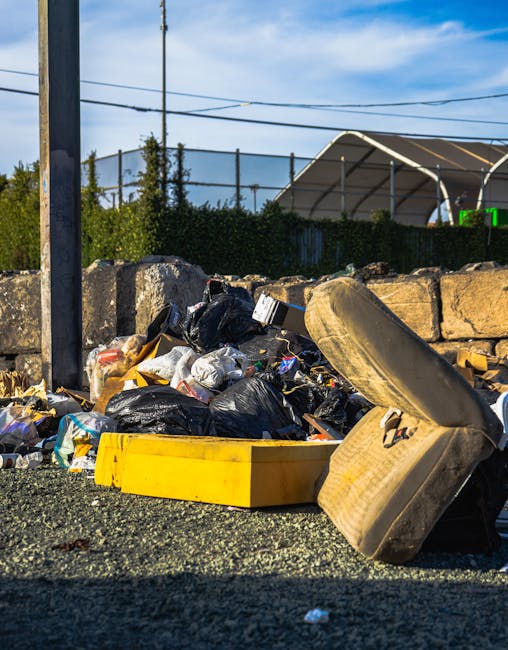 A narrow urban alleyway filled with an accumulation of large black and white garbage bags and cardboard boxes stacked towards the back, with some bags leaning against cluttered wooden pallets. The ground is uneven and covered in dirt, with visible patches of wetness and debris scattered around. To the left, a dark red brick wall features graffiti, while on the right, a grey wall with metal bars and vent fixtures can be seen. Bare tree branches extend overhead, with a glimpse of orange brick buildings in the background behind the pile of waste, indicating a typical city environment that may require private rubbish collection services such as those provided by Rubbish Clearance Waterloo. The scene suggests an area designated for waste disposal, perhaps for on-site clearance, with the makeshift arrangement of rubbish emphasizing the need for proper management and removal, common in settings where residents or small businesses rely on independent waste management solutions rather than local authority collections.
