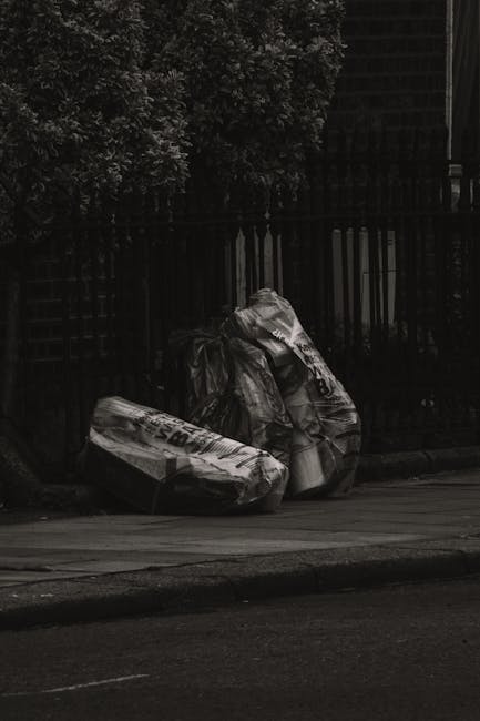 Three large black plastic garbage bags filled with waste are placed on the sidewalk near a wooden fence and a dense leafy bush. The bags appear to be sealed but bulge with contents, indicating they are full. The scene is set outdoors on a street with a paved curb, and the background includes dark fencing and greenery, suggesting a residential area. The lighting in the image is dim, casting shadows on the bags and surroundings, typical of early evening or overcast conditions. The scene illustrates an instance of private rubbish disposal, possibly managed by an independent waste clearance service like Rubbish Clearance Waterloo, where refuse is collected for removal separate from municipal systems. The bags' surfaces show creases and crinkles, and one bag has visible printed markings, hinting at commercial or household waste. Overall, the image showcases typical waste bags ready for collection, emphasizing the importance of proper rubbish handling and disposal in urban environments.