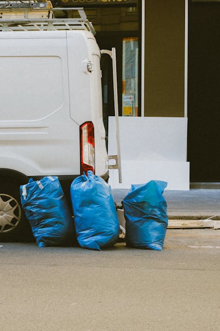 The image shows the rear end of a white commercial van parked on an urban street. Next to the van, on the curb, are three large blue rubbish bags made of plastic, filled and tied at the top, with some appearing slightly wrinkled and sagging. Behind the bags, there is a white structure that looks like a small platform or step, leaning against the building facade, which has large glass windows and a dark-colored wall. The pavement surface is smooth and grey, with a faint division line visible in the foreground. The scene is lit with natural daylight, indicating a clear or partly cloudy day, and the overall setting suggests a typical environment for local rubbish collection or private waste disposal services, such as those offered by Rubbish Clearance Waterloo in South Bank, London, supporting alternative waste handling outside of council services.