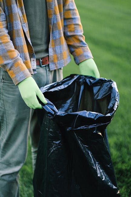 A person standing outdoors on a grassy area, wearing a plaid shirt in shades of yellow, blue, and gray over a dark green top, with green rubber gloves on their hands. They are holding open a large black rubbish bag, which appears to be made of durable plastic, with the opening held wide to facilitate waste collection. The person's trousers are light gray, and a brown belt is visible around the waist. The background features a lawn or garden setting with well-maintained, lush grass. This scene is indicative of waste collection or rubbish removal activities, such as preparing for rubbish clearance or private waste disposal, consistent with services offered by Rubbish Clearance Waterloo for same-day rubbish removal in South Bank and Waterloo areas.
